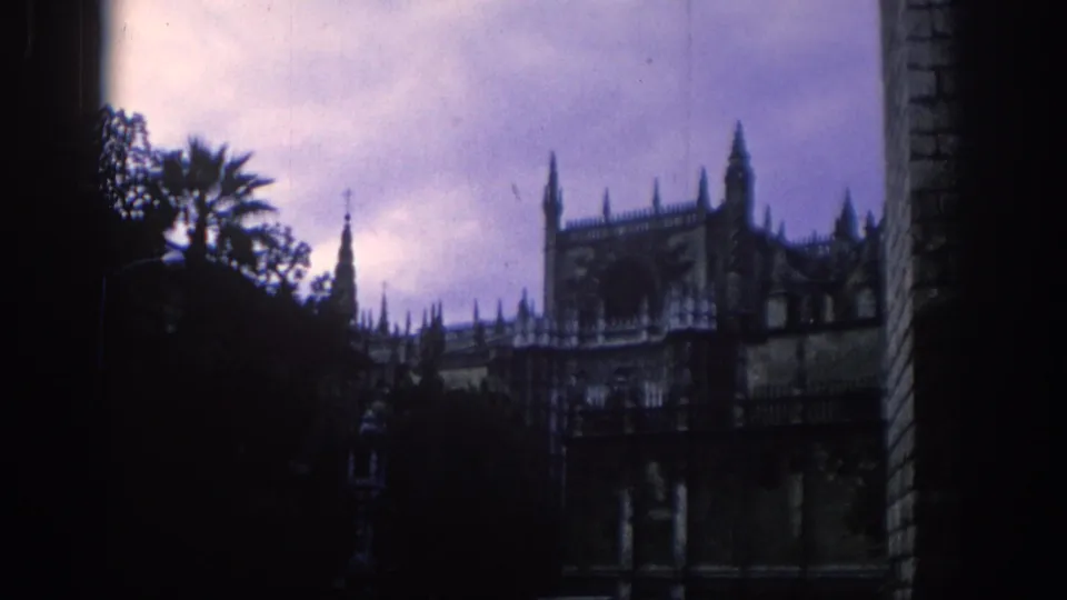 1966 Spain 8mm frame showing a darker wide view of spired architecture, palm foliage, cloudy sky and a vertical stone edge.