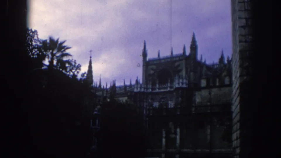 1966 Spain home-movie frame with palm foliage, spired historic architecture, cloudy purple sky and dark 8mm exposure.