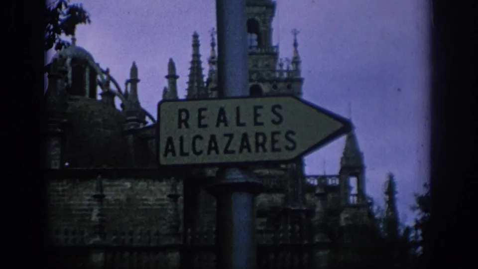 1966 Spain 8mm frame with a Reales Alcazares directional sign against ornate spires and dark foreground edges.