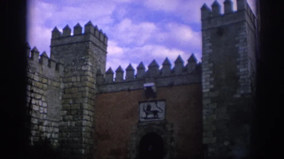 1966 Spain 8mm view of a medieval-looking fortified wall with crenellated towers, an arched doorway, and soft cloudy sky.