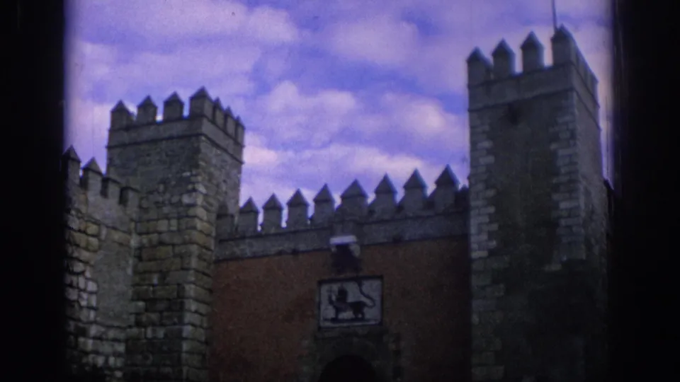 1966 Spain 8mm frame showing a stone fortress facade with crenellated battlements, square towers, and a heraldic lion panel above a reddish gate wall.
