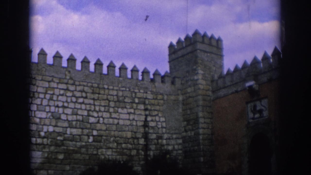 1966 Spain 8mm frame of a crenellated stone wall and tower, with a reddish gate section and heraldic lion panel visible at the right.
