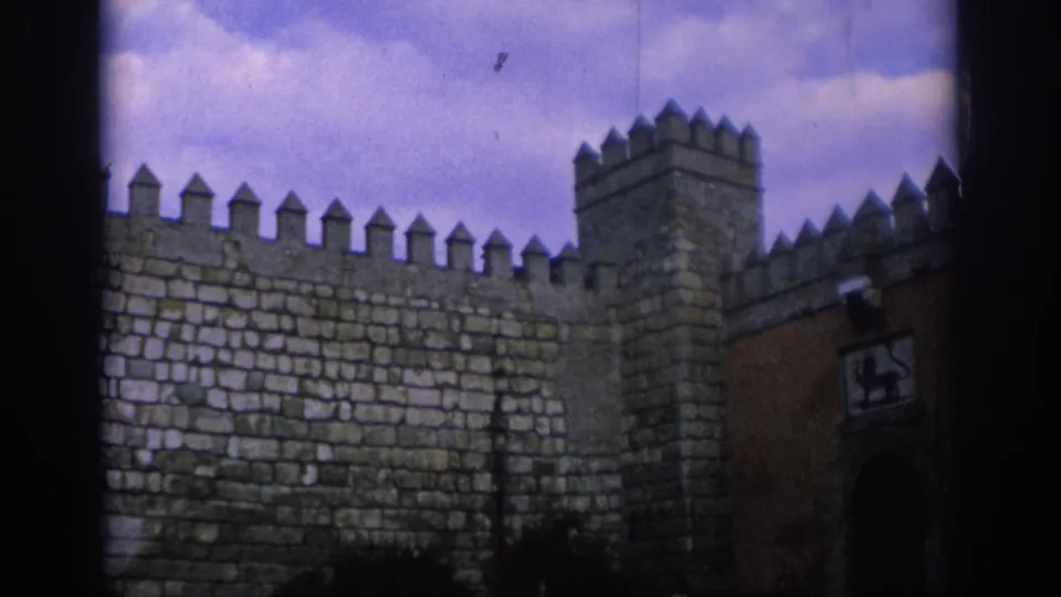 1966 Spain 8mm frame of a crenellated stone wall and tower, with a reddish gate section and heraldic lion panel visible at the right.