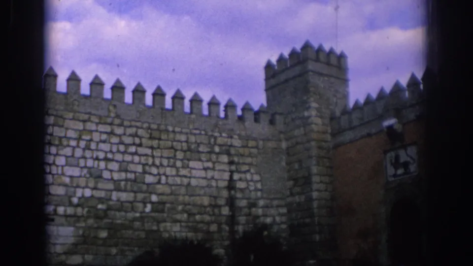 1966 Spain 8mm frame showing a wide fortress wall with crenellations, a square tower, reddish gate masonry, and dark shrub silhouettes below.
