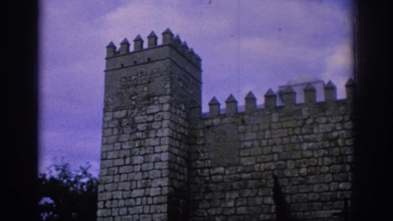 1966 Spain 8mm frame showing a stone castle-like wall with battlements and a square tower under a color-shifted cloudy sky.