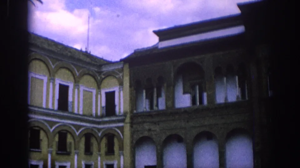 1966 Spain home-movie frame showing a courtyard corner with stacked arcades, arched windows, and a dark roof edge in purple-tinted light.