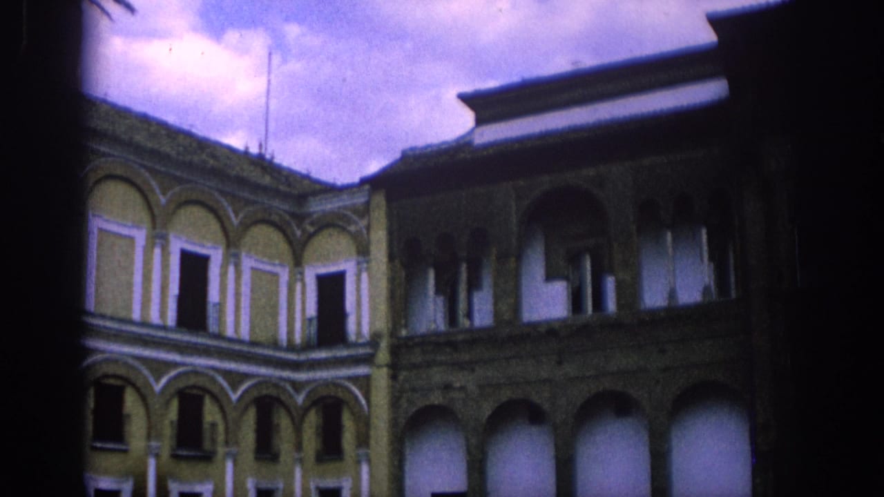 1966 Spain 8mm frame showing a historic courtyard with arcaded walls, upper windows, stone pillars and deep shadow.
