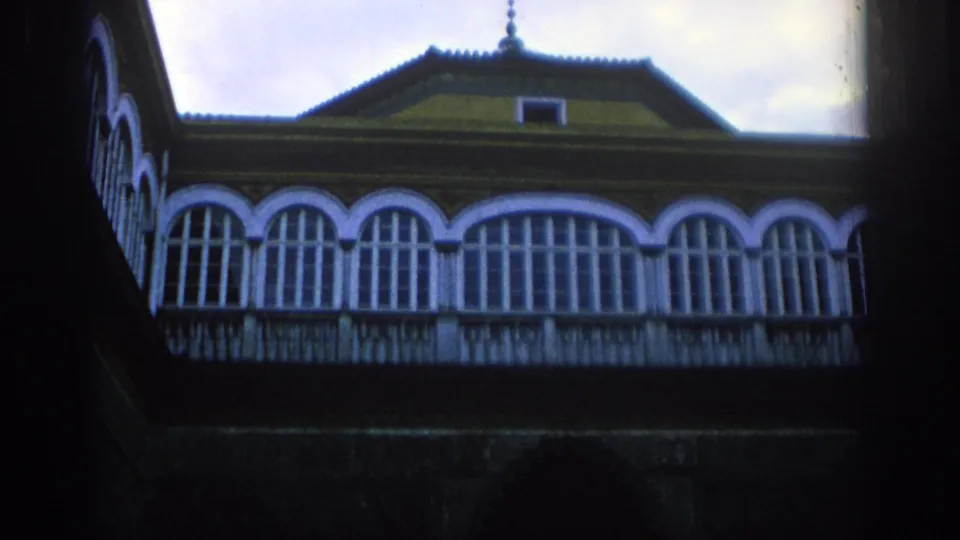 Static 8mm view in Spain, 1966, of a historic facade with repeated arched windows, a balcony rail, and a roofline against pale sky.
