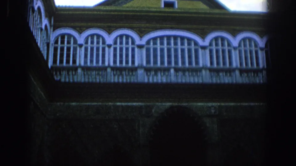 1966 Spain travel footage frame with arched balcony glazing, decorative railing, tiled roof, and a dark lower wall in the foreground.