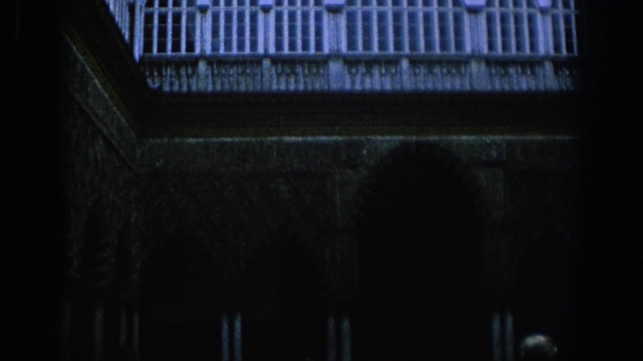 1966 Spain 8mm frame showing a dim historic courtyard with arched columns, balcony railings, and a woman near the lower edge.