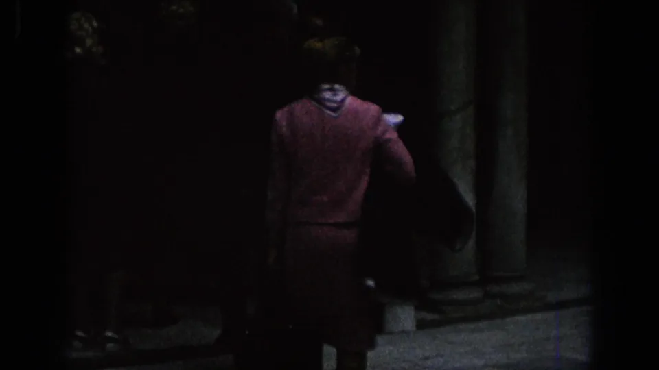 1966 Spain 8mm frame showing a woman from behind near stone columns and a shaded courtyard wall.
