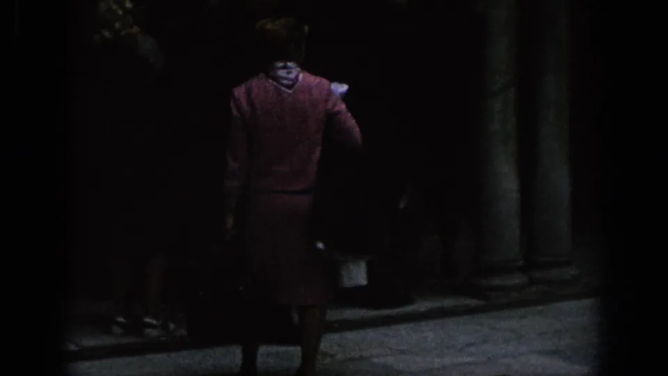 1966 Spain 8mm frame of a woman viewed from behind in a shadowed historic courtyard with columns and dark openings.