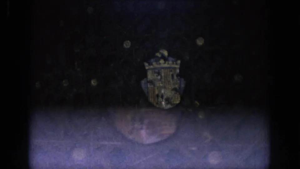 Low-light 8mm frame from Spain showing a crowned shield near the top of a dark surface with a bright exposure band below.