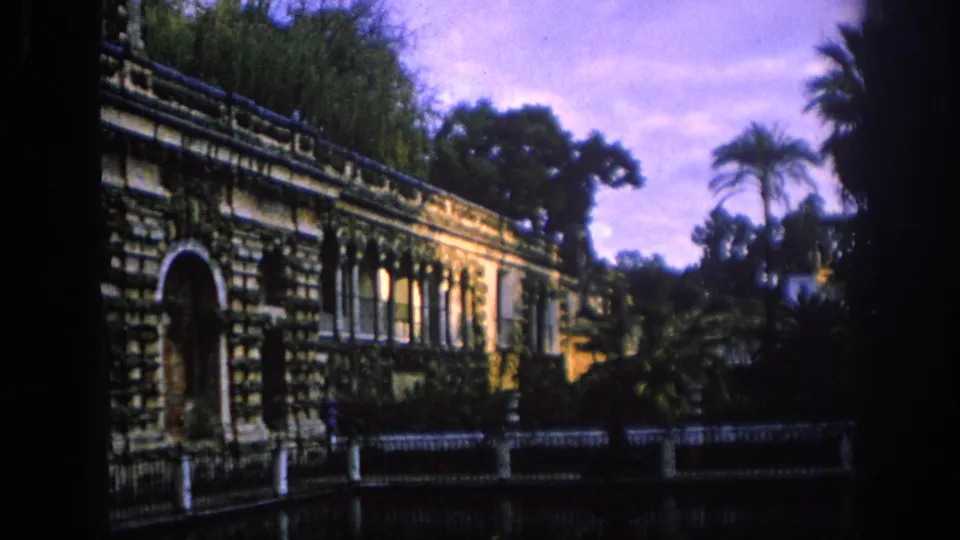 1966 Spain home-movie view of an ornate stone building beside greenery, palms, water, and a purple-blue sky.