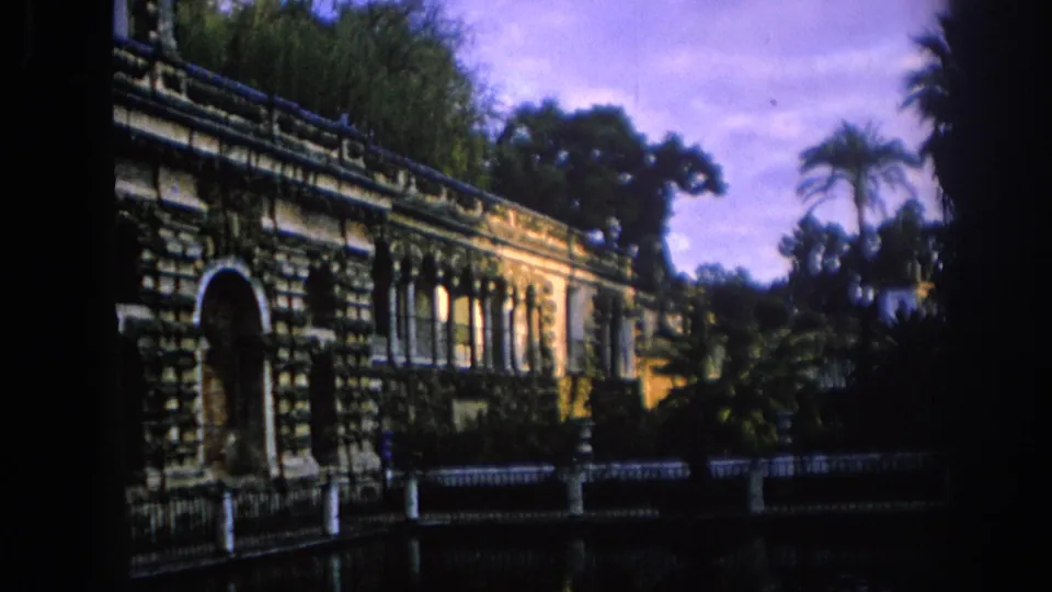 1966 Spain 8mm frame showing a closer visitor-shot view of a stone building with arched entrance, palms, garden plants, and water in front.