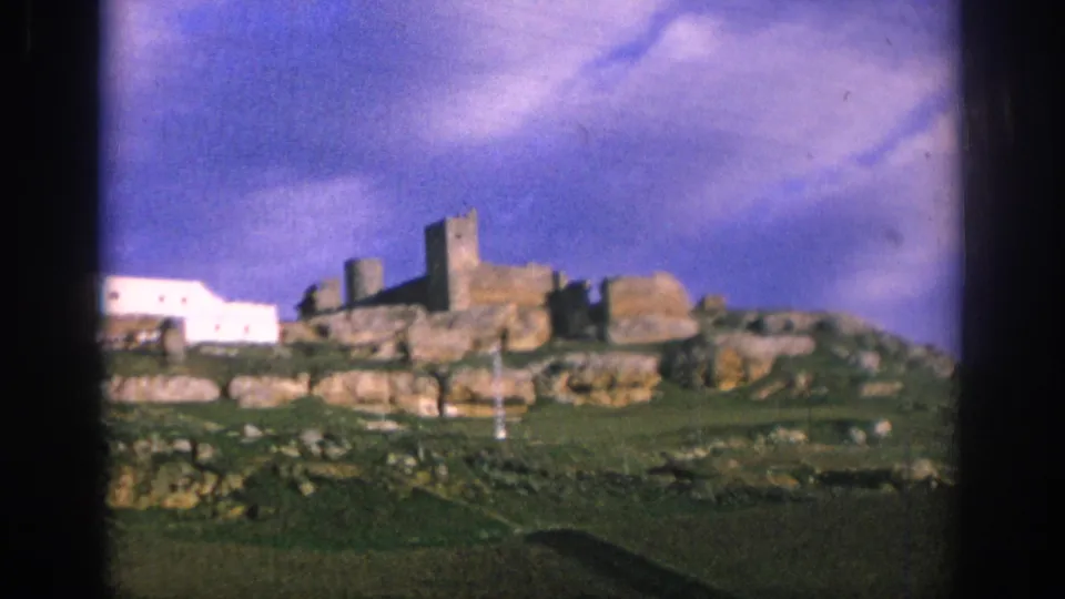 1966 Spain home-movie frame of a distant stone ruin-like structure on rocky green terrain under a blue, clouded sky.