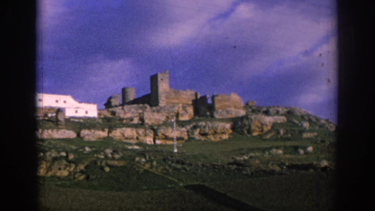 1966 Spain 8mm frame showing distant stone ruins, whitewashed buildings, rocky hillside, and open sky.