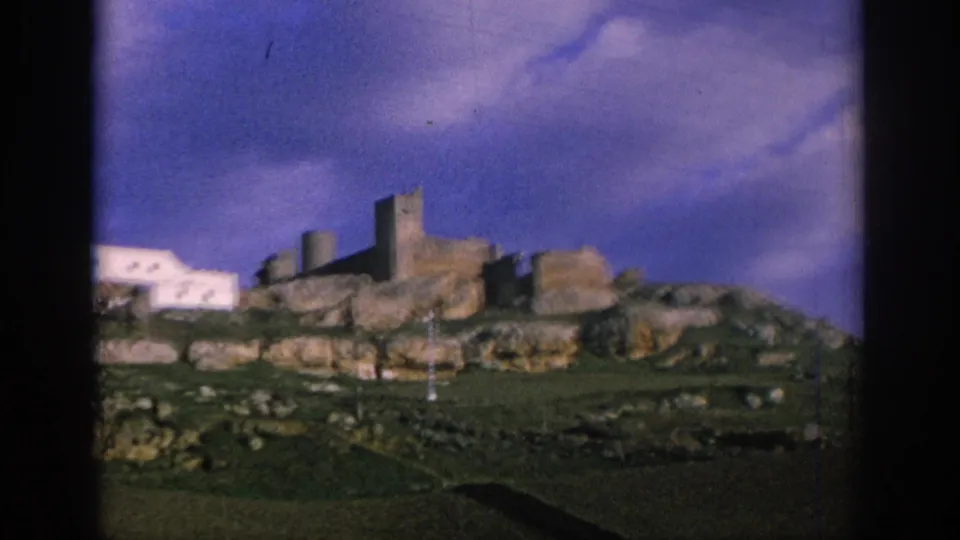1966 Spain travel footage frame with fortress-like ruins, white structures, green hillside, and soft 8mm color shift.