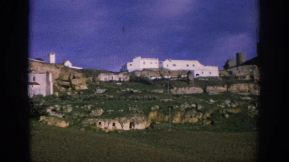 1966 Spain 8mm frame showing white hillside buildings, low stone features, rocky ground, and a dark blue sky.