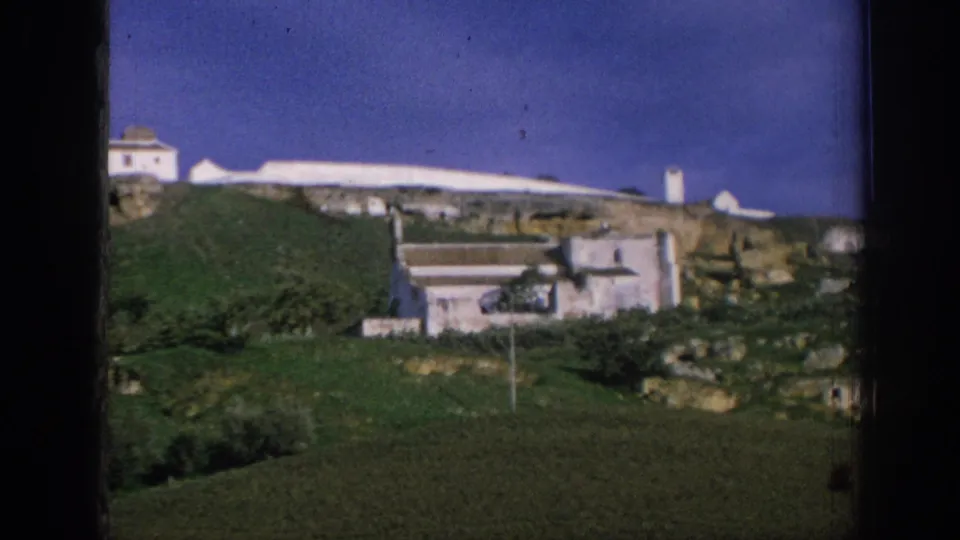 1966 Spain home-movie frame with white buildings, a terraced green hill, rocky ledges, and dark film borders.