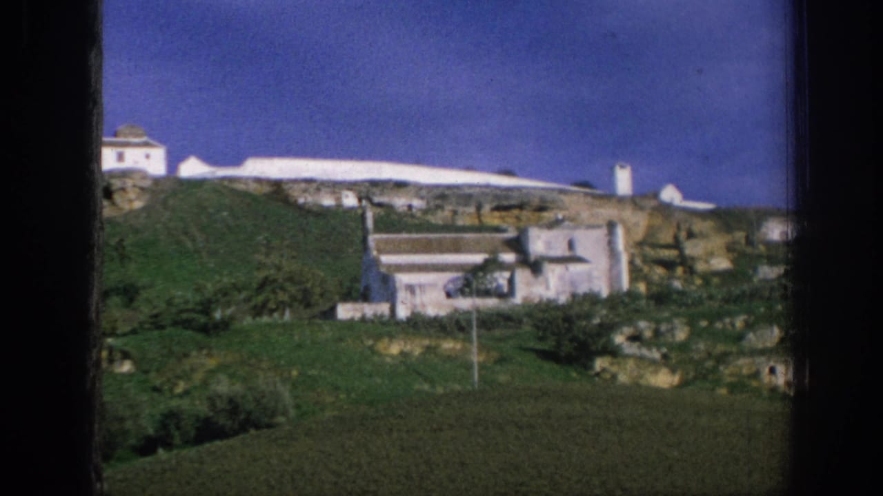 1966 Spain 8mm frame showing whitewashed hillside buildings, terracotta roofs, and green slopes under a saturated blue sky.