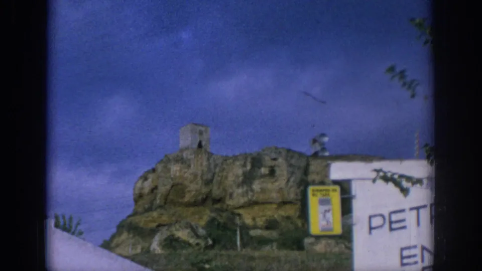 1966 Spain travel frame with a rocky outcrop, small stone tower, white wall, yellow sign case, and partial painted lettering.