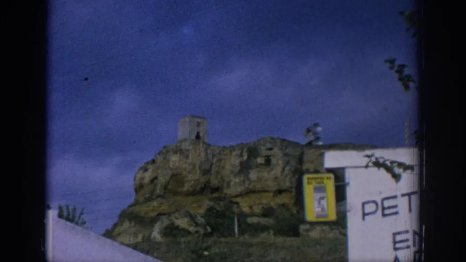 1966 Spain 8mm frame repeating the rocky tower view with a white wall, poster case, partial painted text, and blue sky.