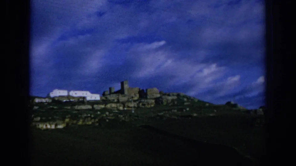 1966 Spain 8mm frame showing distant white buildings and old stone structures along a ridge beneath a vivid blue sky.