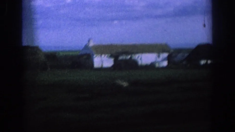 1966 Spain 8mm frame from a moving car, showing a white farmhouse and dark-roofed rural structure beyond fields.