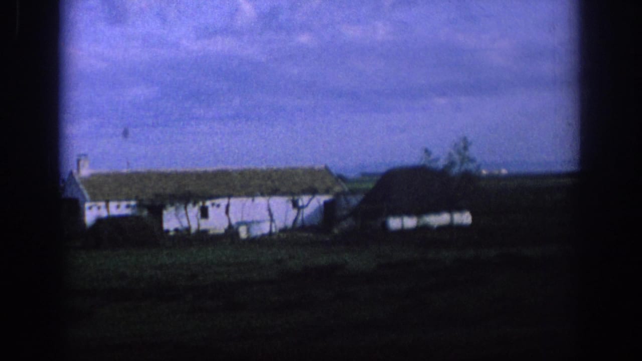 White farmhouse and dark-roofed structure seen through a dark vehicle window frame across open land.