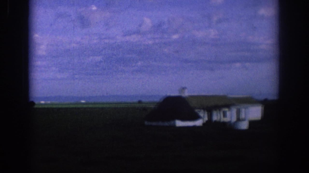 1966 Spain home-movie frame showing a small white farmhouse beside open fields, seen from a moving viewpoint under a tinted cloudy sky.