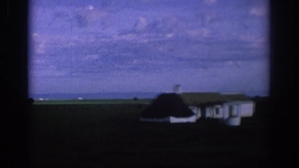 1966 Spain home-movie frame showing a small white farmhouse beside open fields, seen from a moving viewpoint under a tinted cloudy sky.