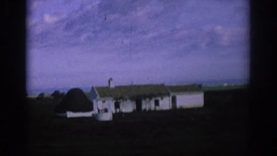 1966 Spain 8mm frame showing a closer moving-vehicle view of a white rural house with attached low buildings and surrounding fields.