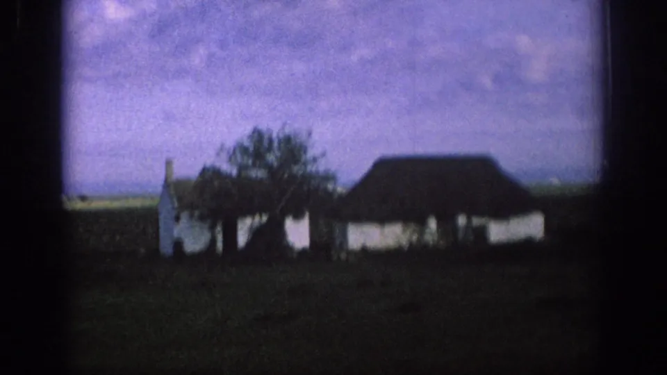 1966 Spain 8mm frame showing a white farmhouse with a front tree, dark roof, shadowed field, and soft purple sky.