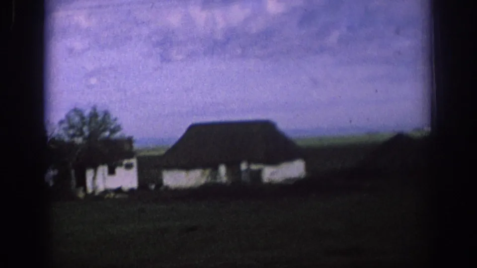 1966 Spain 8mm frame of a white rural building with a dark roof, nearby tree, green field, and low countryside horizon.