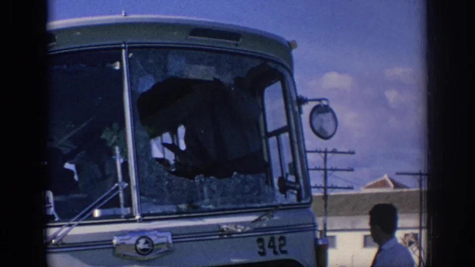 1966 Spain home-movie frame showing the broken bus windshield, side mirror, and a man in a light shirt and tie beside it.