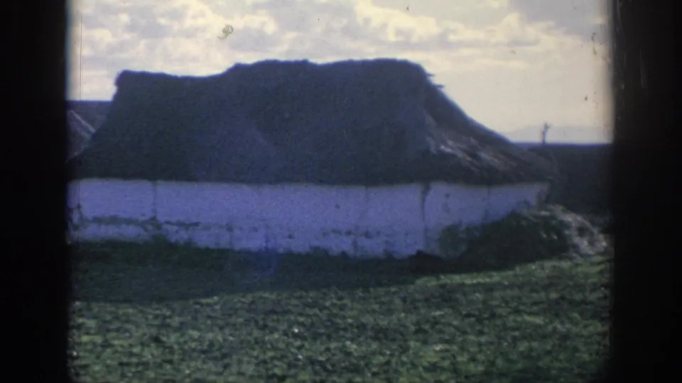 1966 Spain 8mm frame looking toward a whitewashed rural building with a dark thatched roof under a bright sky.