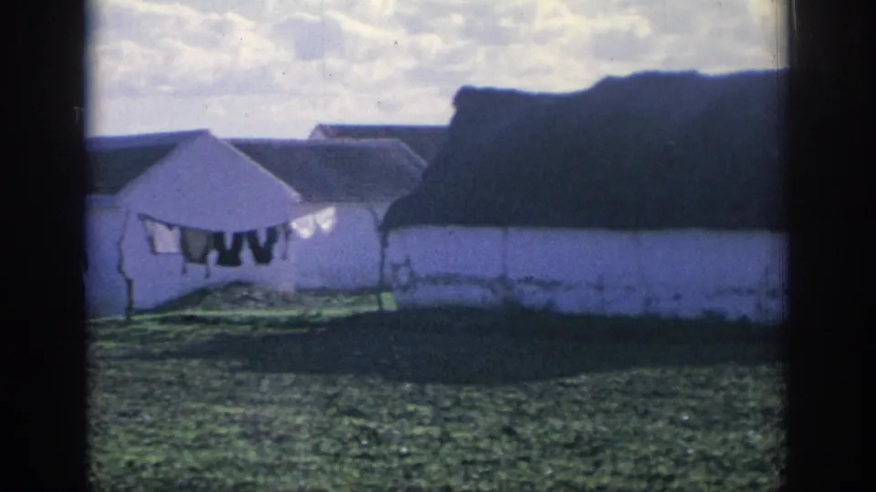1966 Spain 8mm frame showing white rural buildings, dark roofs, a clothesline with laundry, grass, and sky.