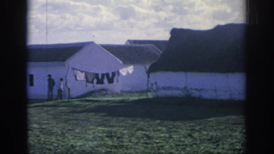 1966 Spain 8mm frame showing whitewashed farm buildings, a laundry line, and distant people in a grassy rural yard.