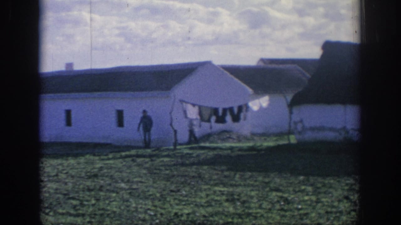 1966 Spain home-movie frame with a white farm structure, laundry hanging outside, and one distant person by the building.