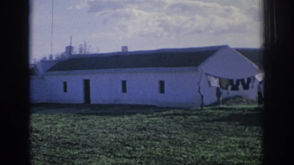 1966 Spain rural home-movie frame with a whitewashed farm building, laundry line, grassy foreground, and cloudy sky.