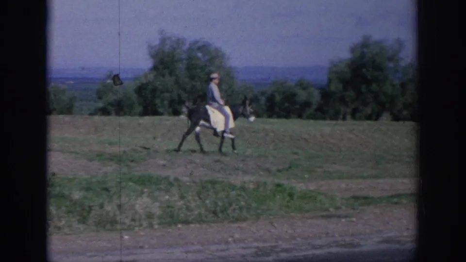 1966 Spain 8mm frame showing a man in light clothing riding a donkey across an open field under a hazy sky.
