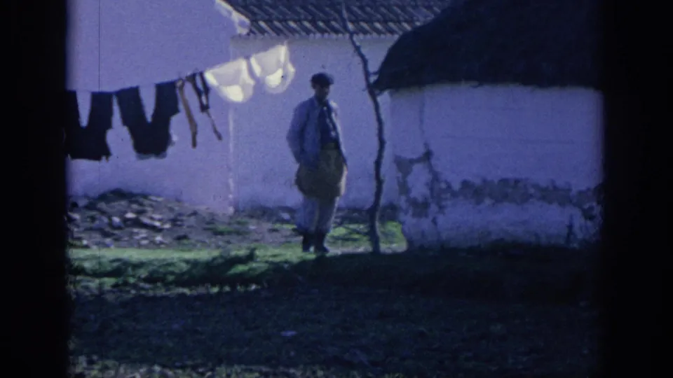 1966 Spain frame showing a man near a whitewashed rural building, outdoor laundry, rough ground, and shadowed roof structure.