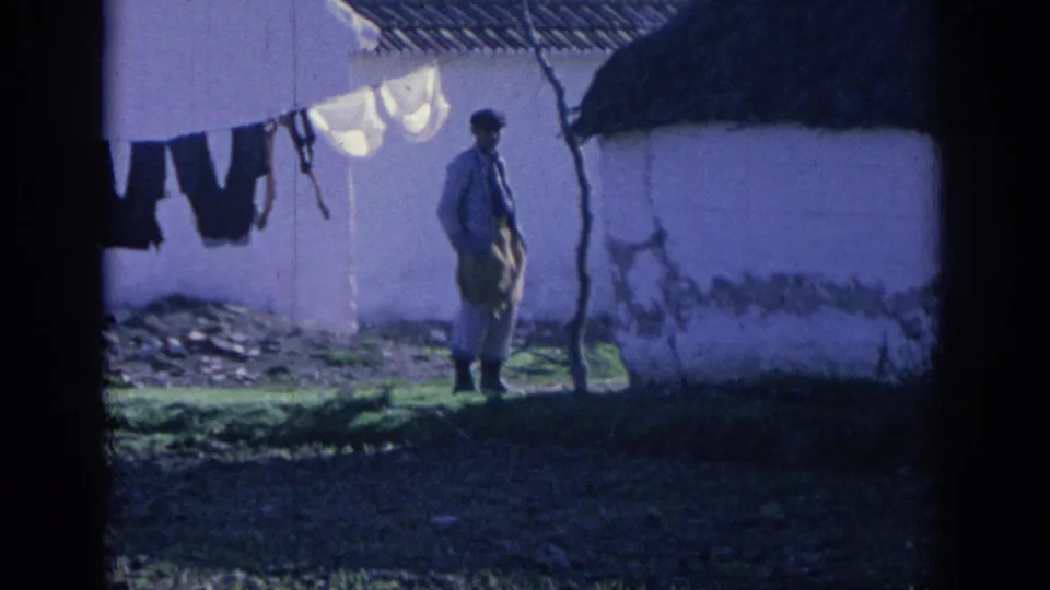1966 Spain home-movie frame of a man holding a sack or bundle near a whitewashed village building and hanging laundry.