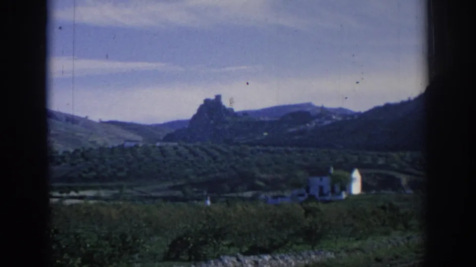 1966 Spain 8mm countryside frame showing valley fields, hillside contours, small rural buildings and distant rocky high ground.