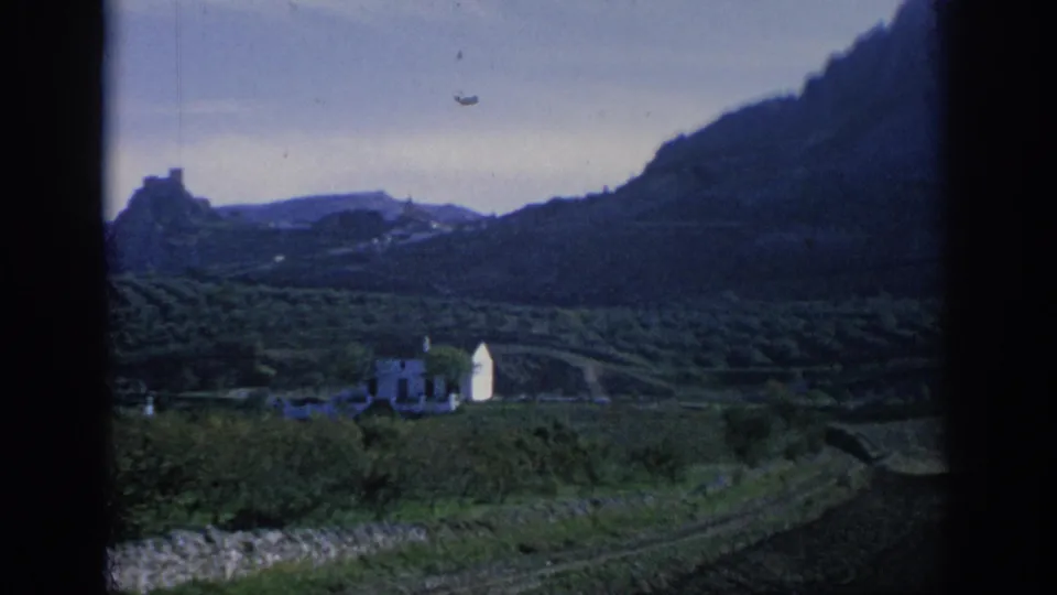 1966 Spain 8mm frame showing terraced green hills, a white valley building, distant rocky high ground, and dark window-frame edges.
