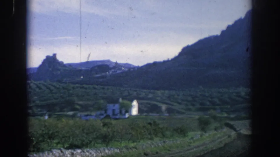 1966 Spain 8mm frame with a white building below terraced hills and a distant hilltop fort-like structure near a mountain slope.