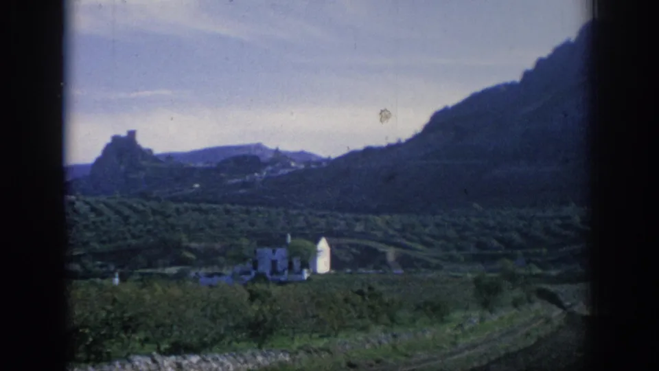 1966 Spain 8mm frame showing a terraced rural valley, white building, distant rocky landmark, and soft blue-green film color.