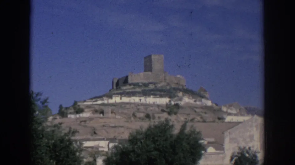 1966 Spain 8mm frame showing a stone hilltop fort above white buildings, dry slope, blue sky, and foreground vegetation.
