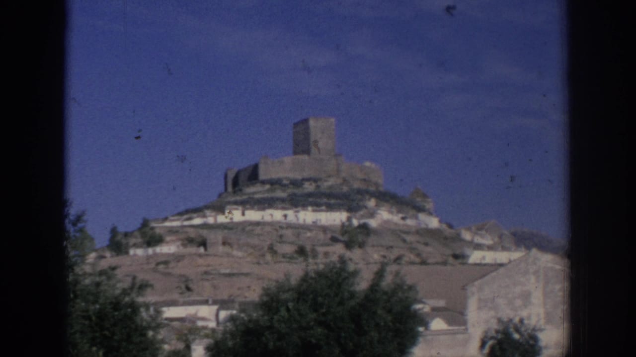 Hilltop stone castle above white buildings and trees under a blue sky.